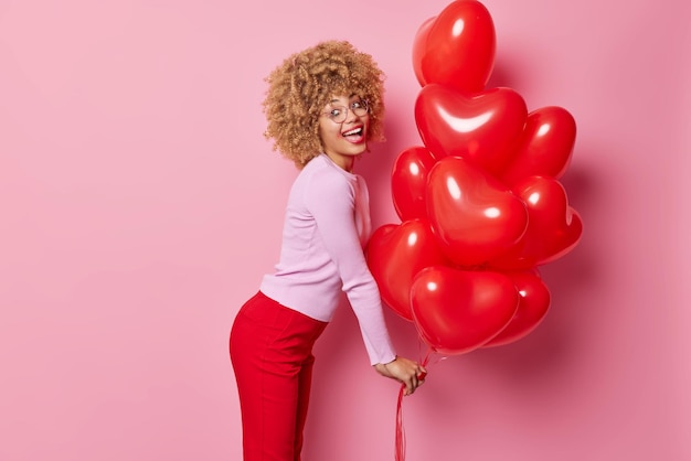 Fröhliche lockige junge Europäerin trägt Pullover und rote Hosen steht seitlich und hält Heliumherzballons mit durchgesickertem Make-up. Romantische Stimmung fühlt sich energisch in Posen vor rosafarbenem Hintergrund an