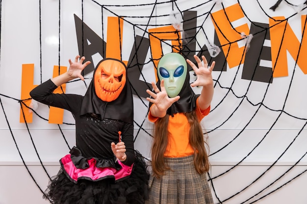 Fröhliche mädchen, die vor der halloween-partydekoration stehen und kürbis jacko'lantern und alien-maske tragen