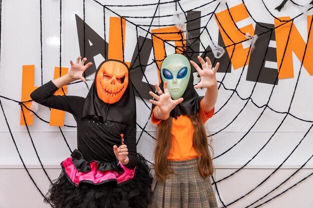 Fröhliche Mädchen, die vor der Halloween-Partydekoration stehen und Kürbis Jacko'lantern und Alien-Maske tragen