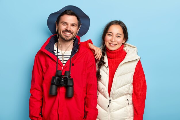 Fröhliche Mischlinge Frau und Mann lächeln freudig, haben Freizeit, in lässigem Outfit gekleidet, benutzen ein Fernglas für die Expedition, posieren an der blauen Wand