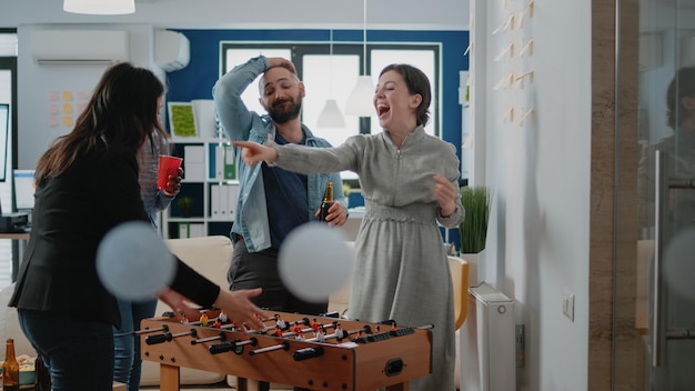 Fröhliche Mitarbeiter, die nach der Arbeit ein Fußballspiel am Tischfußball genießen. Kollegen spielen und trinken Getränke, um die Party im Büro zu feiern. Frau, die Fußballspiel gewinnt und Spaß hat.