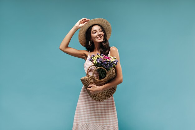 Fröhliche moderne Brünette mit Ohrringen und coolem Hut im stilvollen leichten Kleid posiert mit Strohhandtasche und bunten Blumen