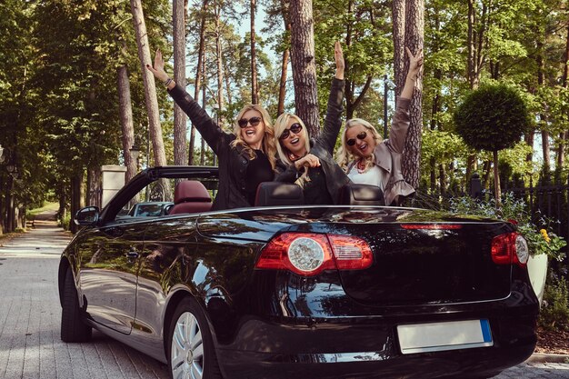 Fröhliche modische Freundinnen heben ihre Hände, während sie in einem Luxus-Cabriolet-Auto in einem Park sitzen.