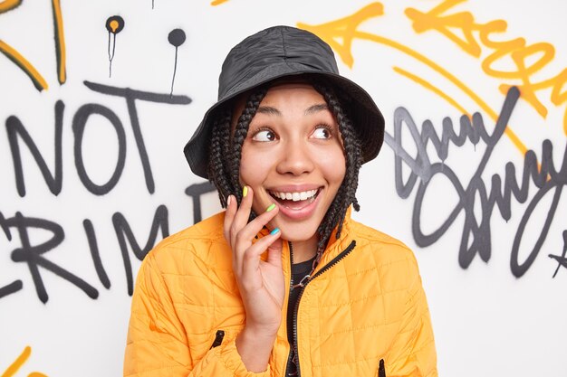 Fröhliche modische junge Frau lächelt glücklich schaut beiseite trägt stilvolle Kleidung verbringt Freizeit in städtischen Ort posiert vor bunten Graffiti-Wand