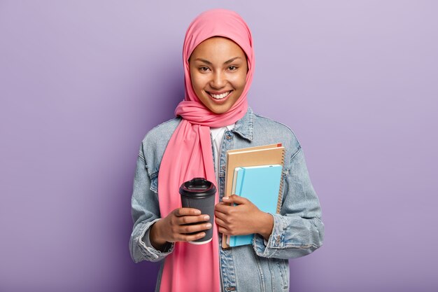 Fröhliche muslimische Frau in rosa Hijab, Jeansmantel, trägt Taschenbuch