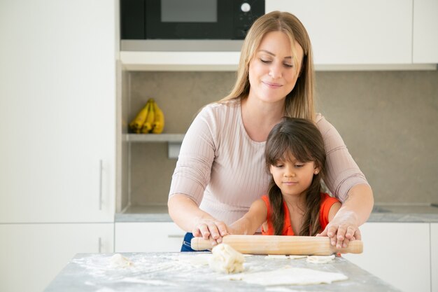 Fröhliche Mutter und ihr Mädchen kochen zusammen und rollen Teig auf Küchentisch mit Mehlpulver.