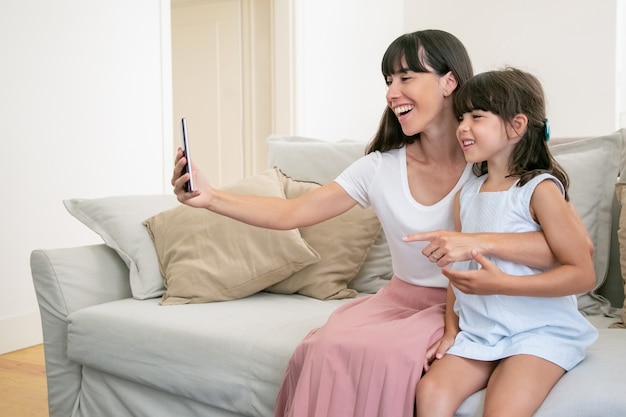 Fröhliche Mutter und kleine Tochter, die Telefon für Videoanruf verwenden, während sie zusammen auf Sofa zu Hause sitzen