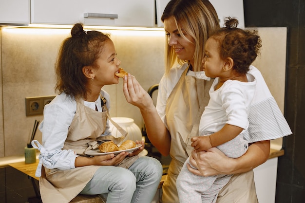 Fröhliche Mutter und kleine Tochter essen frisch gebackene Kekse in der Küche, genießen hausgemachtes Gebäck, tragen Schürzen und lächeln einander an, haben Spaß zu Hause. Mutter, die ihr Tochterkleinkind hält