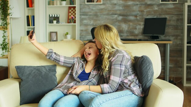 Fröhliche Mutter und Tochter machen ein Selfie mit Smartphone im Wohnzimmer.