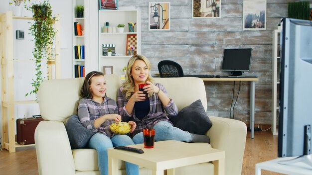 Fröhliche Mutter und Tochter sitzen auf der Couch im Wohnzimmer und schauen fern, essen Chips und trinken Soda.