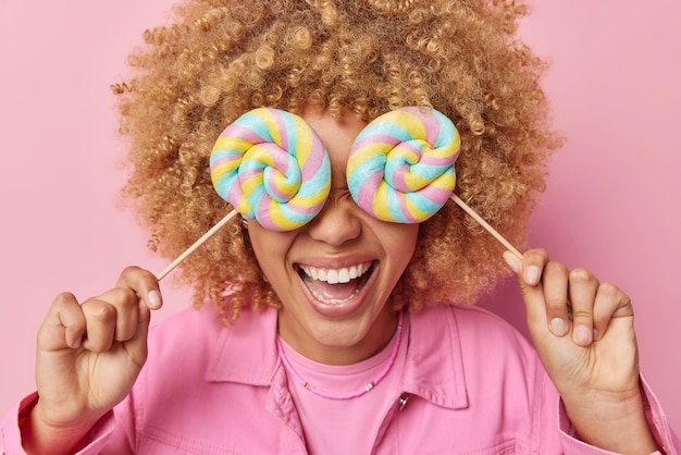 Fröhliche, nicht erkennbare lockige Frau bedeckt die Augen mit süßen Lollipops auf Stöcken hat Zuckersucht genießt Cheat Meal Day in legerer Kleidung Posen vor rosa Hintergrund Schädliches Essen