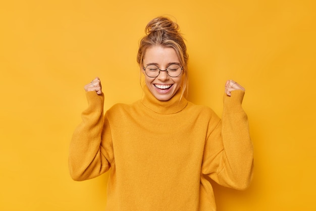 Kostenloses Foto fröhliche, positive frau mit haaren, die zu einem knoten zusammengeballt sind, freut sich über den erfolg, feiert den sieg oder ein großartiges ergebnis, trägt einen lässigen pullover, runde brille, posiert vor einer gelben wand. ja, ich habe es endlich geschafft