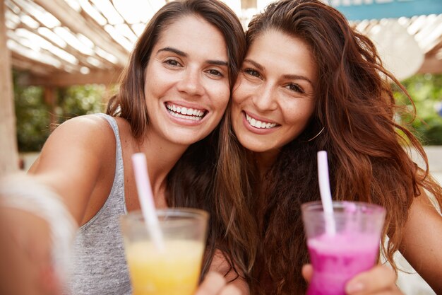 Fröhliche positive Frauen posieren vor der Kamera eines nicht wiedererkennbaren Geräts, machen Selfie als Getränke mit frischen, farbenfrohen Cocktails und sitzen vor dem Innenraum eines Cafés im Freien.