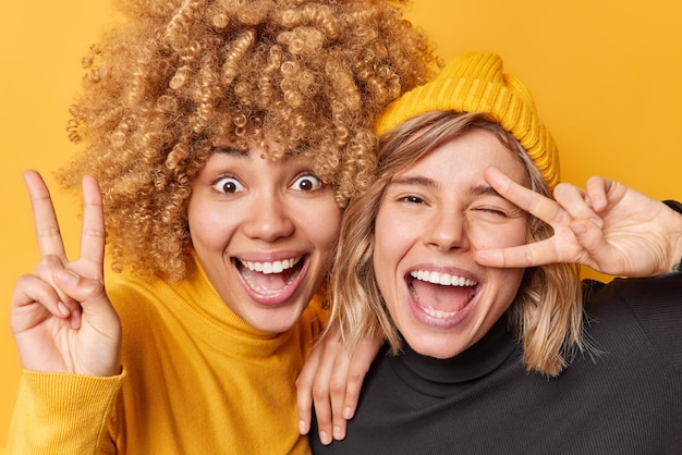 Kostenloses Foto fröhliche positive junge frauen zeigen friedensgeste v-zeichen und schreien positiv gekleidet in freizeitkleidung viel spaß pose indoor vor leuchtend gelben hintergrund siegessymbol körpersprache konzept