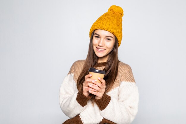 Fröhliche positive Koffeinfrau, Frau in einem Pullover und einem Hut mit Kaffee in ihren Händen, die auf einer weißen Wand aufwerfen