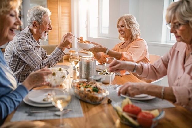 Fröhliche reife Frau, die ihrem Mann Essen übergibt, während sie mit ihren Freunden am Esstisch zu Hause zu Mittag isst