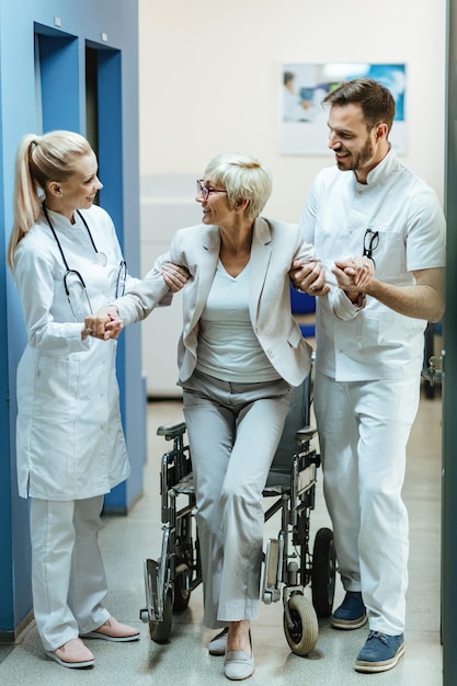 Fröhliche reife Frau, die mit Hilfe von zwei Mitarbeitern des Gesundheitswesens im Krankenhausflur aus einem Rollstuhl aufsteht