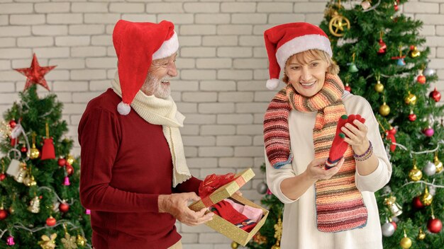 Fröhliche reife frau, die neue handschuhe untersucht, während sie geschenke mit einem älteren ehemann gegen weihnachtsbäume und ziegelmauer austauscht