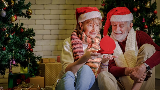 Fröhliche reife frau in weihnachtsmütze, die box öffnet und einem fröhlichen älteren mann ein geschenk zeigt, während sie zusammen gegen weihnachtsbäume und eine mauer sitzt