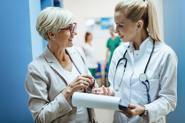 Fröhliche reife Frau und ihr Arzt kommunizieren, während sie in einem Krankenhausflur Papierkram durchgehen