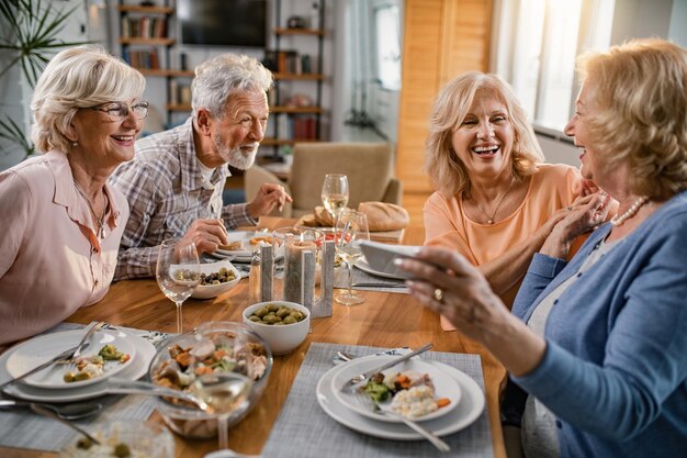 Fröhliche reife Freunde, die zusammen zu Mittag essen und Selfie mit dem Handy am Esstisch machen