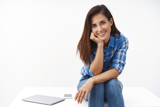 Fröhliche reizende Frau mittleren Alters sitzt am Tisch und hat eine Pause von der Arbeit. Mageres Gesicht, Palme, geneigter Kopf, lächelnd, enthusiastisches Ende, Projekt, Stopp mit Laptop, Smartphone, sprechender Mitarbeiter, weißer Hintergrund