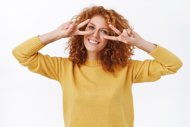 Fröhliche, reizende, lächelnde, rothaarige, lockige Frau im gelben Pullover drückt Positivität und Freude aus, die Zeichen des Friedenssieges über den Augen zeigt, tanzende Disco, kippenden Kopf und grinsende positive Ausdrücke