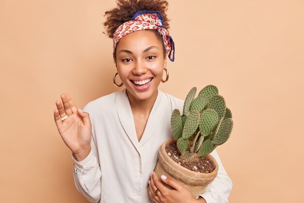 Fröhliche schöne Afro-Amerikanerin hält saftigen Kaktus in Topf lächelt im Großen und Ganzen genießt guten Tag hält die Handfläche angehoben trägt weißes Hemd Kopftuch über den Kopf gebunden isoliert über beige Wand