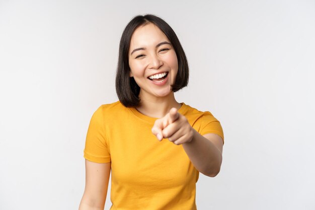 Fröhliche, schöne asiatische Frau, die lachend mit dem Finger auf die Kamera zeigt und kichert, lächelnd, sorglos im gelben T-Shirt auf weißem Hintergrund