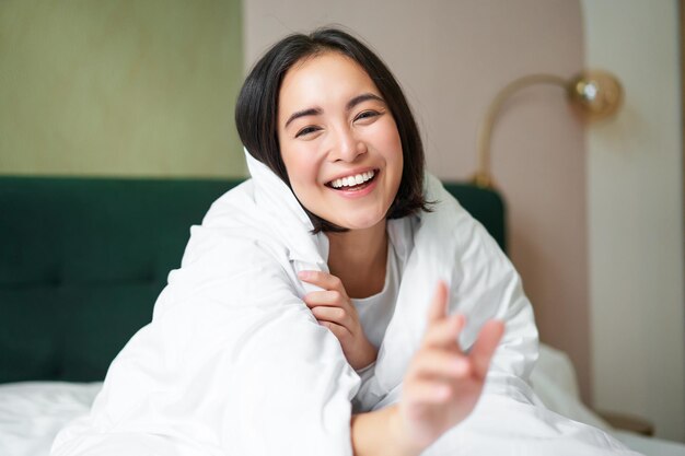 Fröhliche schöne asiatische Frau, die mit einer Decke bedeckt ist, sitzt auf dem Bett in einer warmen Bettdecke und versucht, den Witz der Kamera zu erreichen