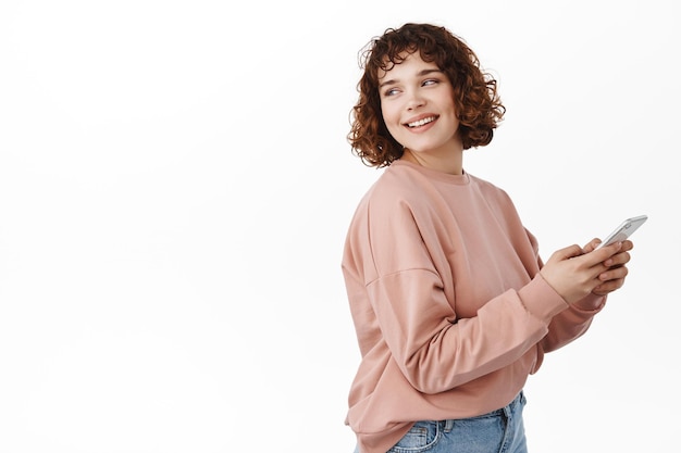 Fröhliche schöne Frau dreht sich mit zufriedenem Lächeln um, nutzt die Chat-App, hält das Smartphone in der Hand und betrachtet den leeren Raum für das Logo-Banner, das vor weißem Hintergrund steht.