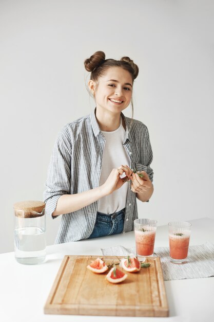 Fröhliche schöne Frau lächelnd, die Grapefruit-Detox-Smoothie mit Rosmarin über weißer Wand verziert. Gesunde Ernährung.