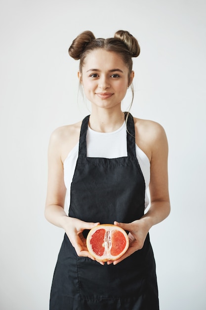 Fröhliche schöne frau mit brötchen in der schürze lächelnd, die hälfte der grapefruit in den händen über weißer wand hält.