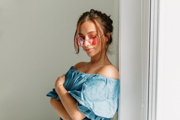 Fröhliche, schöne Frau mit dunklem Haar, rosafarbener runder Brille, blauem T-Shirt mit nackten Schultern, die im Sonnenlicht in der Nähe des Fensters posiert