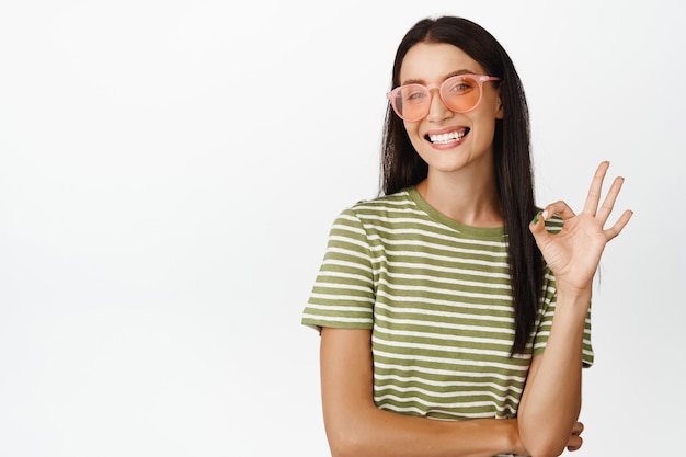 Fröhliche schöne Frau mit stylischer Sonnenbrille zeigt eine okay-Null-Geste und lächelt zufrieden im T-Shirt auf weißem Hintergrund