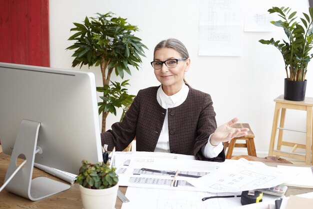 Fröhliche schöne grauhaarige Architektin mittleren Alters, die eine Brille trägt, die lächelt und gestikuliert, während sie vor dem Computer sitzt und sich glücklich fühlt, als sie mit der Arbeit an einem großen Projekt fertig ist