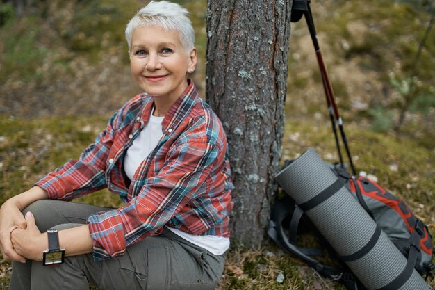 Fröhliche schöne Rentnerin, die unter Baum mit Rucksack und Schlafmatte sitzt und sich während ihrer Reise in der wilden Natur entspannt. Attraktive reife Frau, die Ruhe beim Wandern im Wald hat