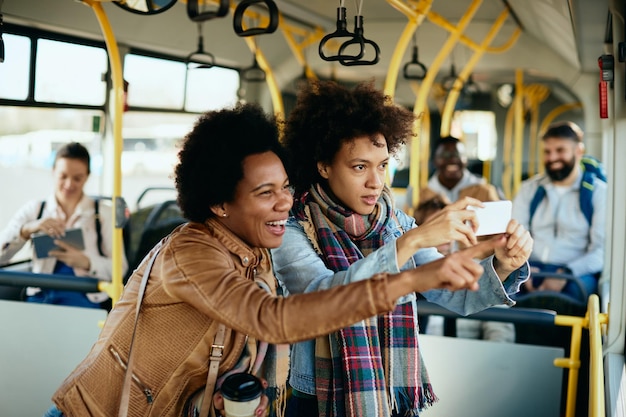 Fröhliche schwarze Freundinnen mit Smartphone und Spaß beim Busfahren