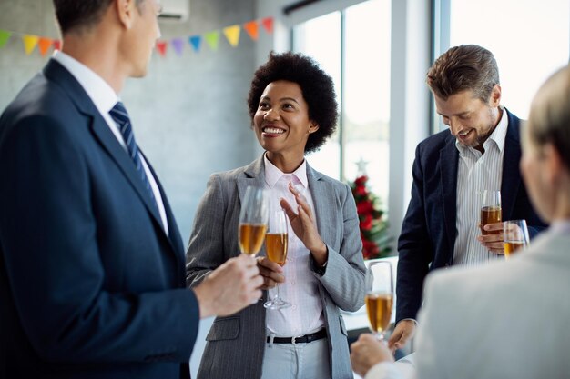 Fröhliche schwarze Geschäftsfrau, die mit Kollegen spricht, während sie auf einer Büroparty Champagner trinkt