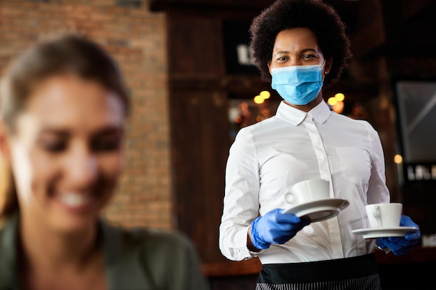 Fröhliche schwarze Kellnerin mit schützender Gesichtsmaske und Handschuhen, die Kaffee in einem Café serviert