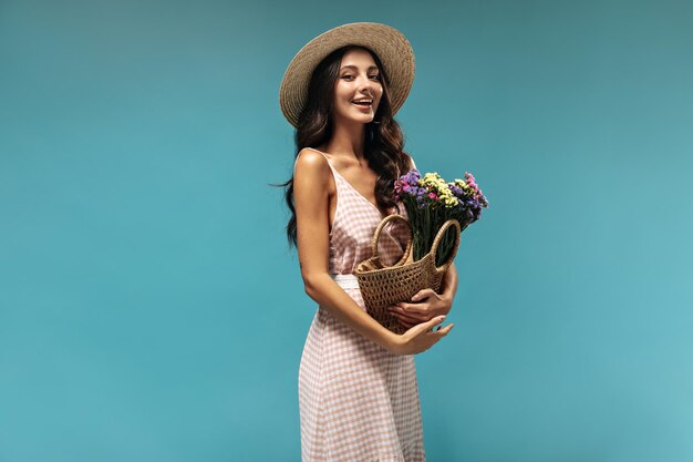 Fröhliche schwarzhaarige Dame mit breitkrempigem Strohhut und langem rosa-weißem Sommerkleid, die lächelt und einen schönen Blumenstrauß hält