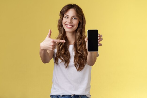 Fröhliche, selbstbewusste, gut aussehende Frau, die ein Foto-Smartphone-Display zeigt, das Handy mit ausgestrecktem Arm Kamera zeigt, die Zeigefinger-Handy-Bildschirm lächelt, erfreut, um die App-Internetanwendung zu fördern
