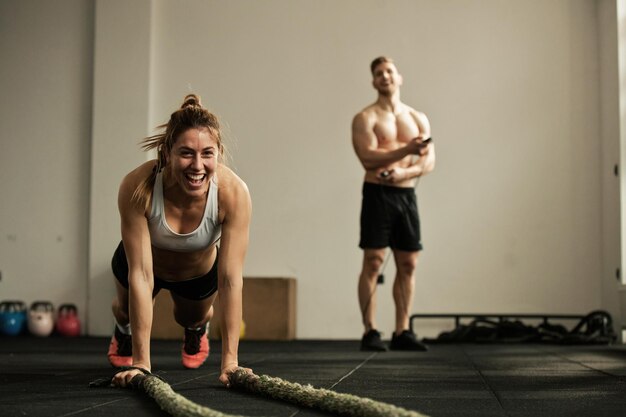 Fröhliche Sportlerin, die Liegestütze macht, während sie mit Kampfseil trainiert und Spaß in einem Fitnessstudio hat. Im Hintergrund ist ein Mann