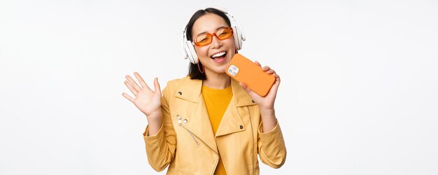 Fröhliche, stilvolle asiatische Frau mit Sonnenbrille, die Musik in Kopfhörern hört und das Smartphone singt und tanzt, das über weißem Hintergrund steht