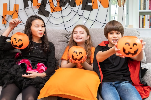 Fröhliche süße Kinder Jungen und Mädchen im Kostüm auf dem Sofa im Wohnzimmer während der Halloween-Party mit Kürbis Jacko'lantern in der Hand