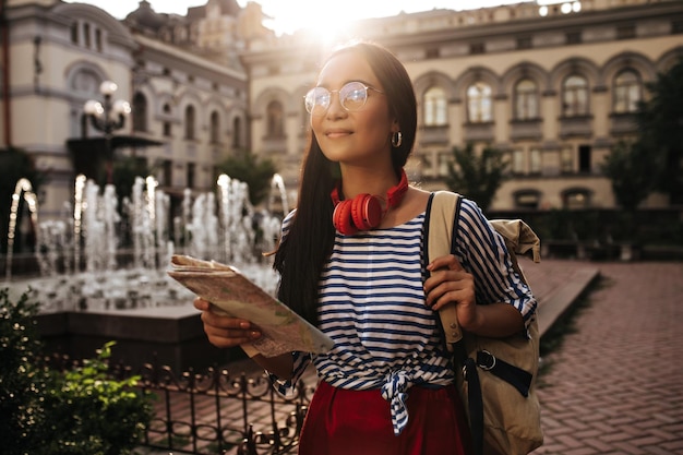 Fröhliche Touristin mit Brille und roten Kopfhörern schaut weg, lächelt, hält Karte und beigen Rucksack draußen