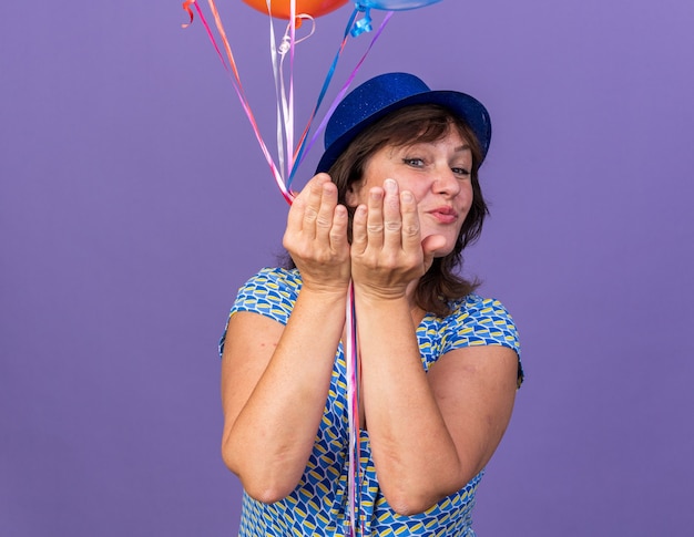 Kostenloses Foto fröhliche und fröhliche frau mittleren alters mit partyhut, die einen haufen bunter ballons hält und einen kuss bläst, um die geburtstagsfeier zu feiern, die über lila wand steht