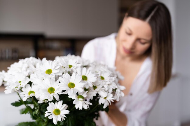 Fröhliche und fröhliche junge Frau in Weiß, die zu Hause in der Küche weiße Blumen arrangiert