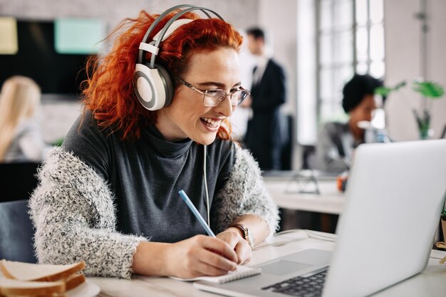 Fröhliche Unternehmerin, die an einem Computer arbeitet und sich Notizen in ihrem Notizblock macht, während sie im Büro Musik über Kopfhörer hört Es gibt Menschen im Hintergrund