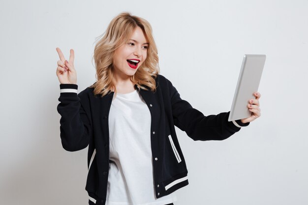 Fröhliche verspielte junge Frau, die Tablet-Computer hält und Friedensgeste zeigt